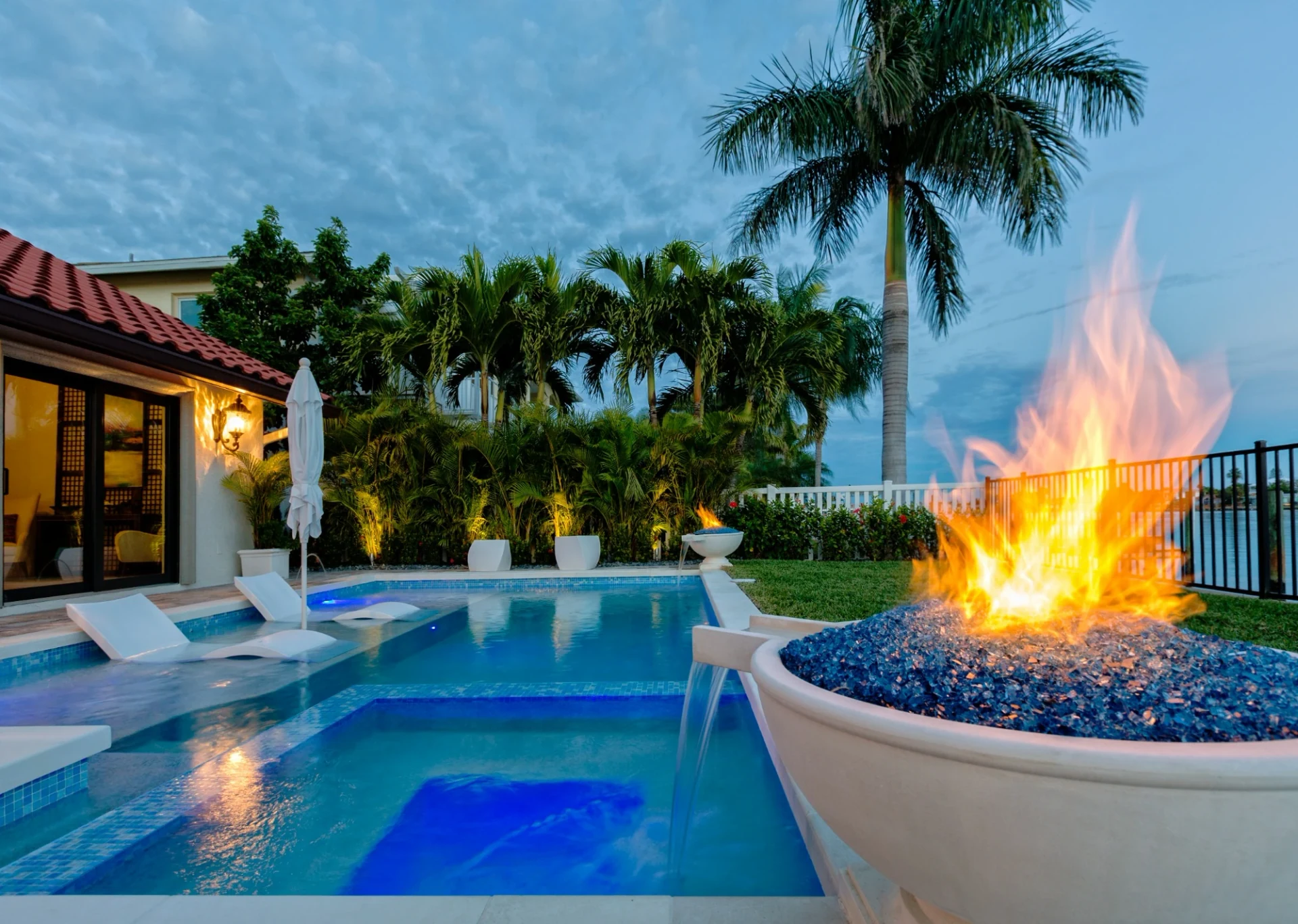 Twilight poolside with a large lit fire bowl, sheer-descent waterfall, uplit palms, and a row of fire pots along the lawn edge