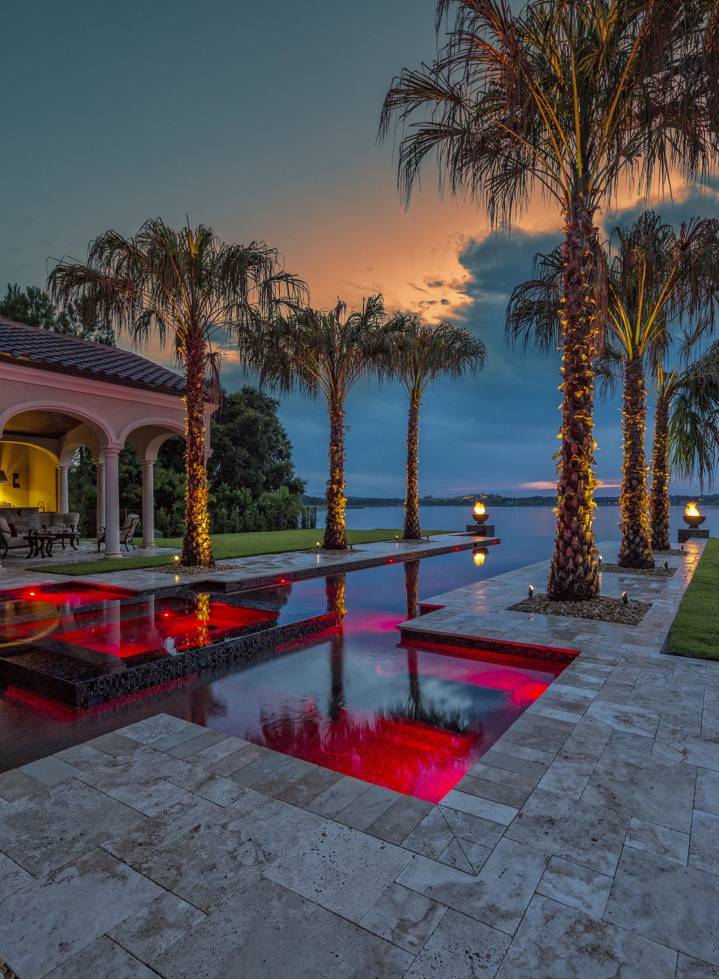 Twilight pool with red-lit water features, uplit royal palms, and an orange-to-blue sunset sky at a luxury Treasure Coast estate.