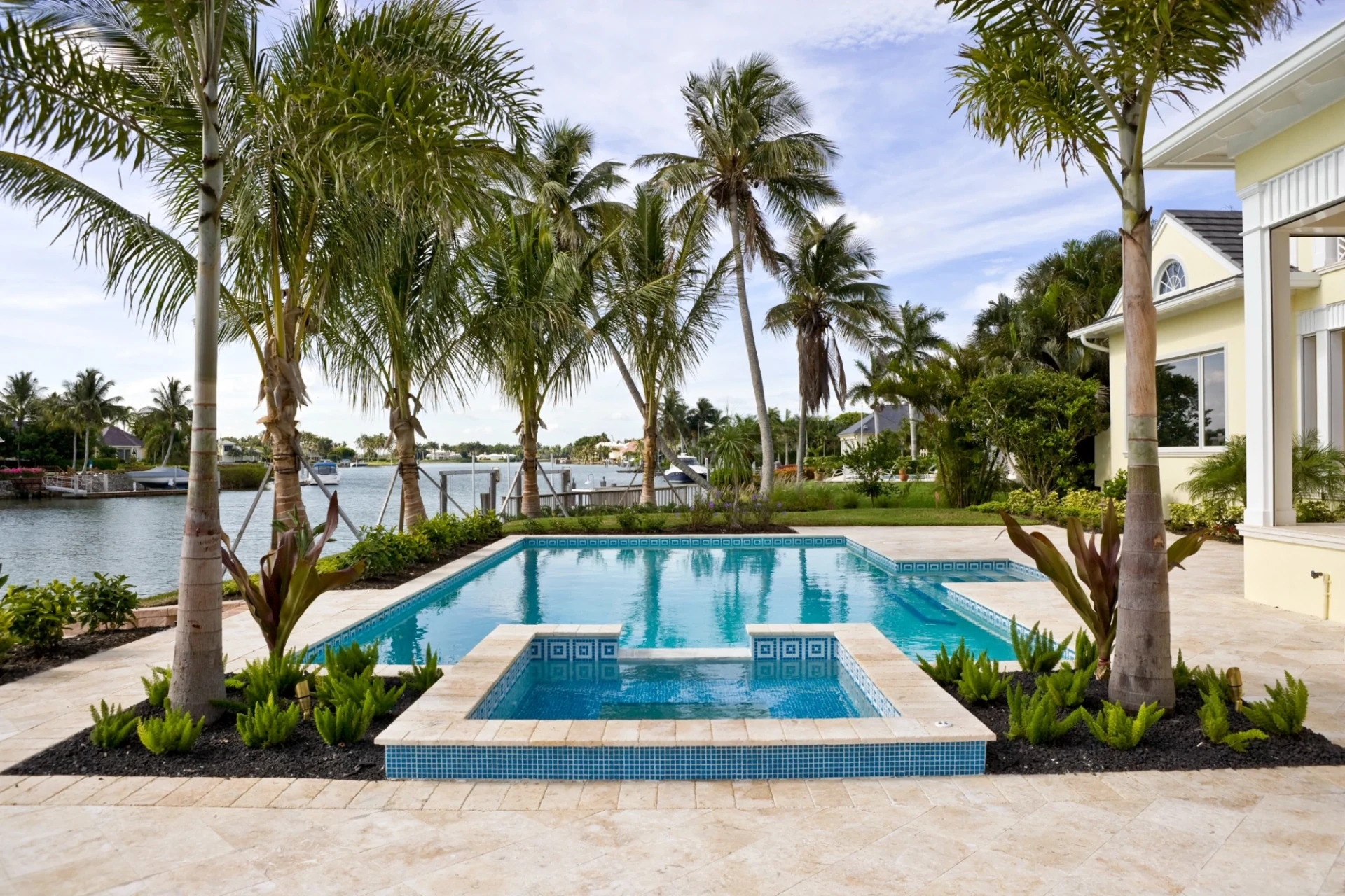 Bright waterfront backyard with a turquoise rectangular pool, raised square mosaic-tile spa, royal palms, light-cream residence to the right, and a canal with docked boats beyond