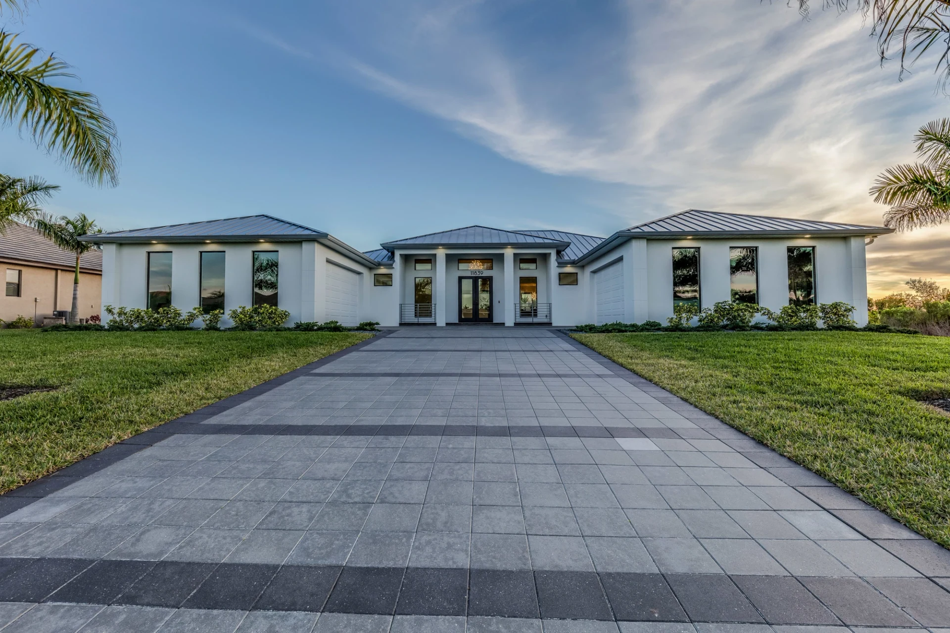 Modern white two-story residence with a long gray paver driveway leading straight to the front entrance, lawn on both sides, royal palms, and a dark standing-seam metal roof under golden-hour light